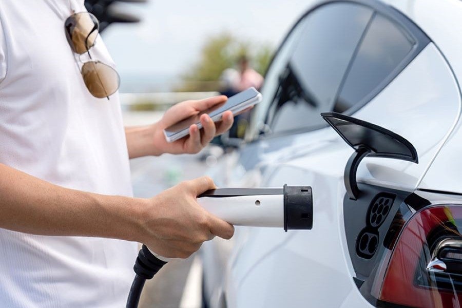 A person holds a Ev charger in one hand and mobile in one hand near white car
